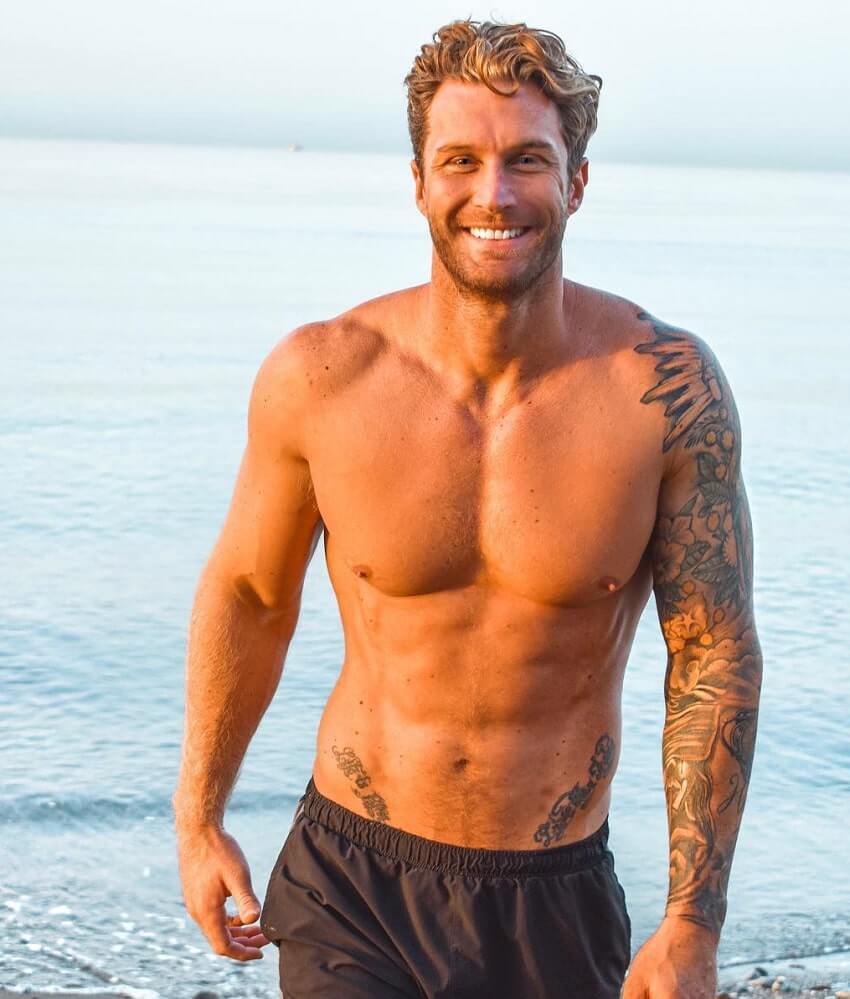 Matt Fox standing shirtless on the beach smiling for the photo, looking fit and lean