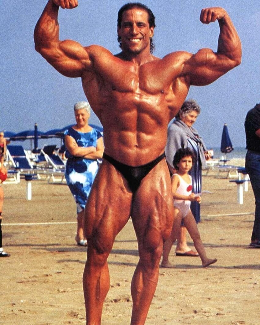 Mike Quinn doing a shirtless front double biceps flex on the beach, looking huge and ripped