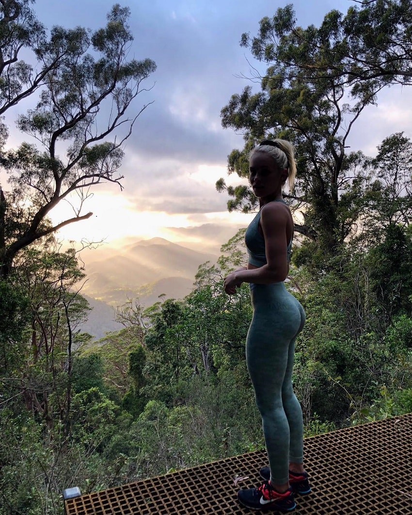 Morgan Rose Moroney standing on the mountain peak among trees and green lush nature, watching a misty sunrise