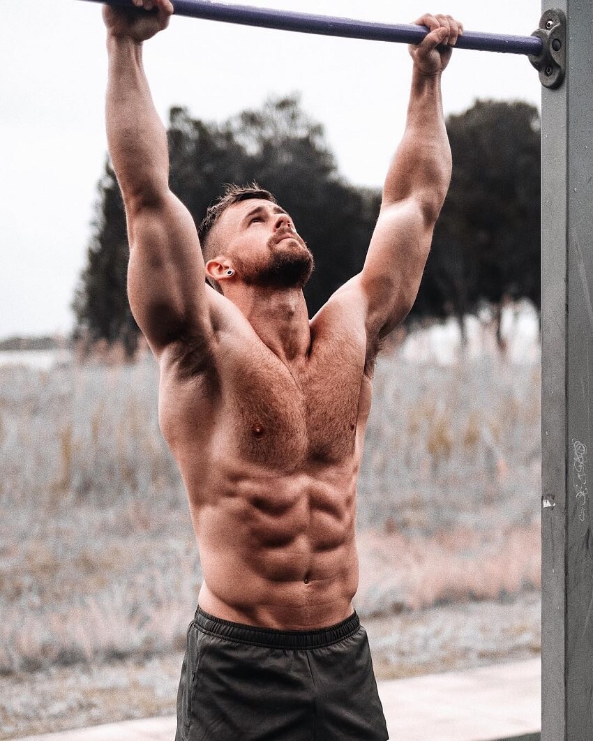 Nathan McCallum reaching for a pull-up bar with his hands while being shirtless, looking ripped and muscular