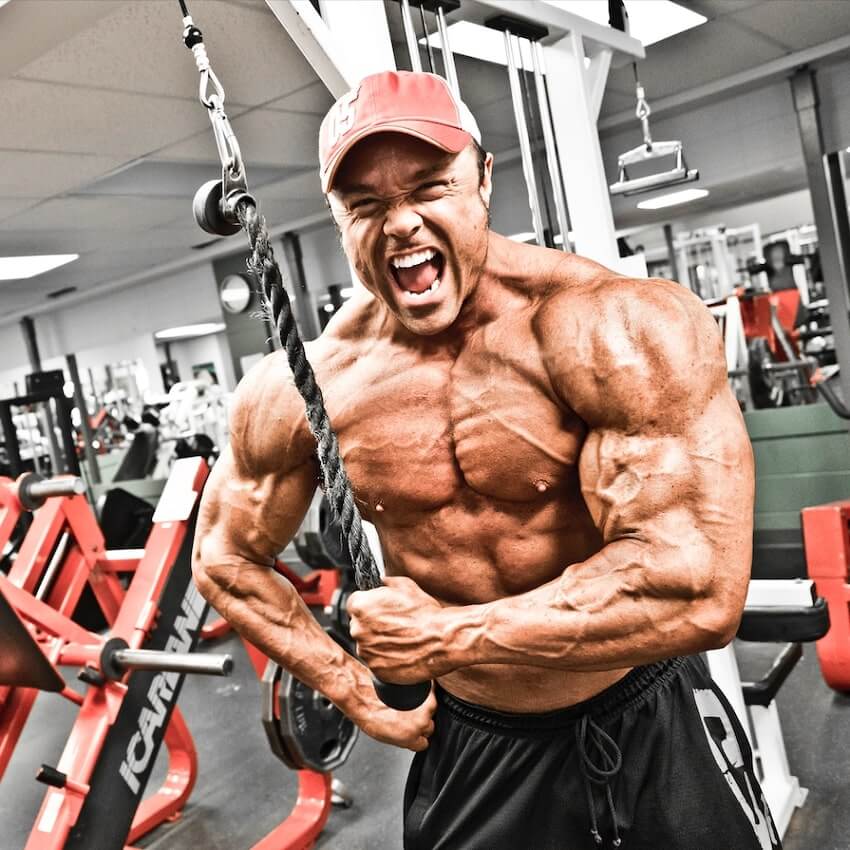 Ron Partlow flexing in the gym while doing triceps extensions, looking huge and ripped