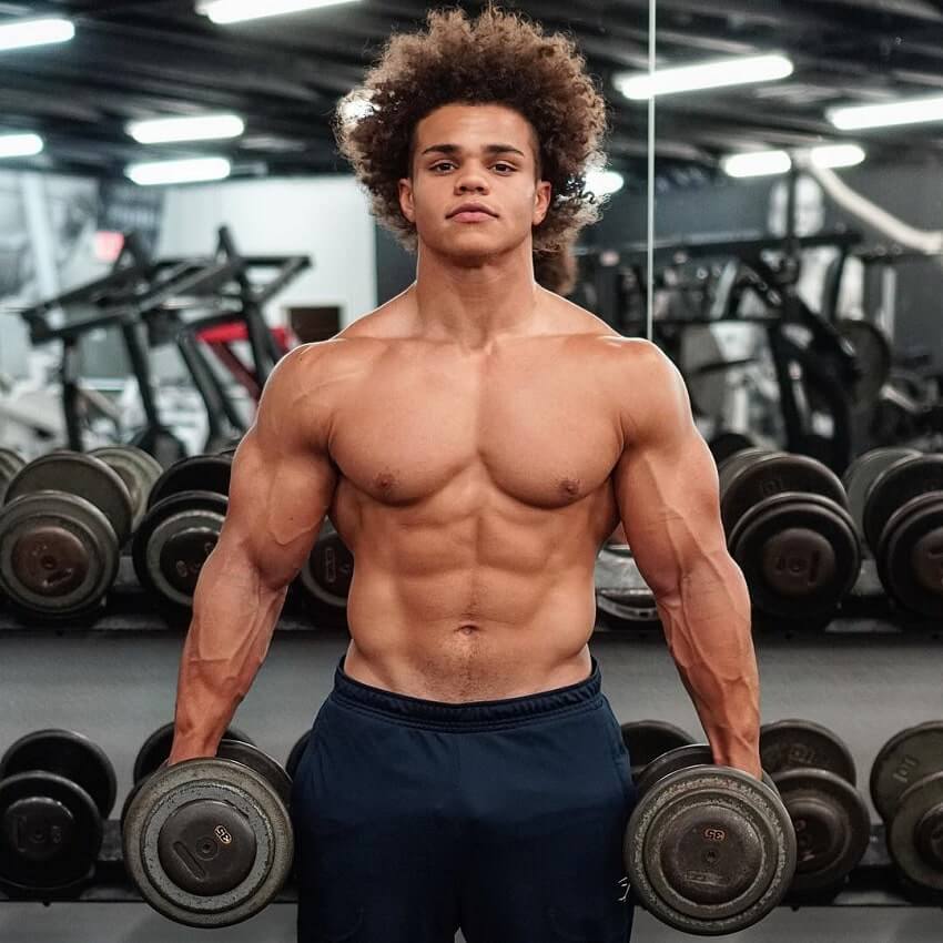 Uzoma Obilor holding two dumbbells in his hand as he poses shirtless for the camera, looking swole and ripped