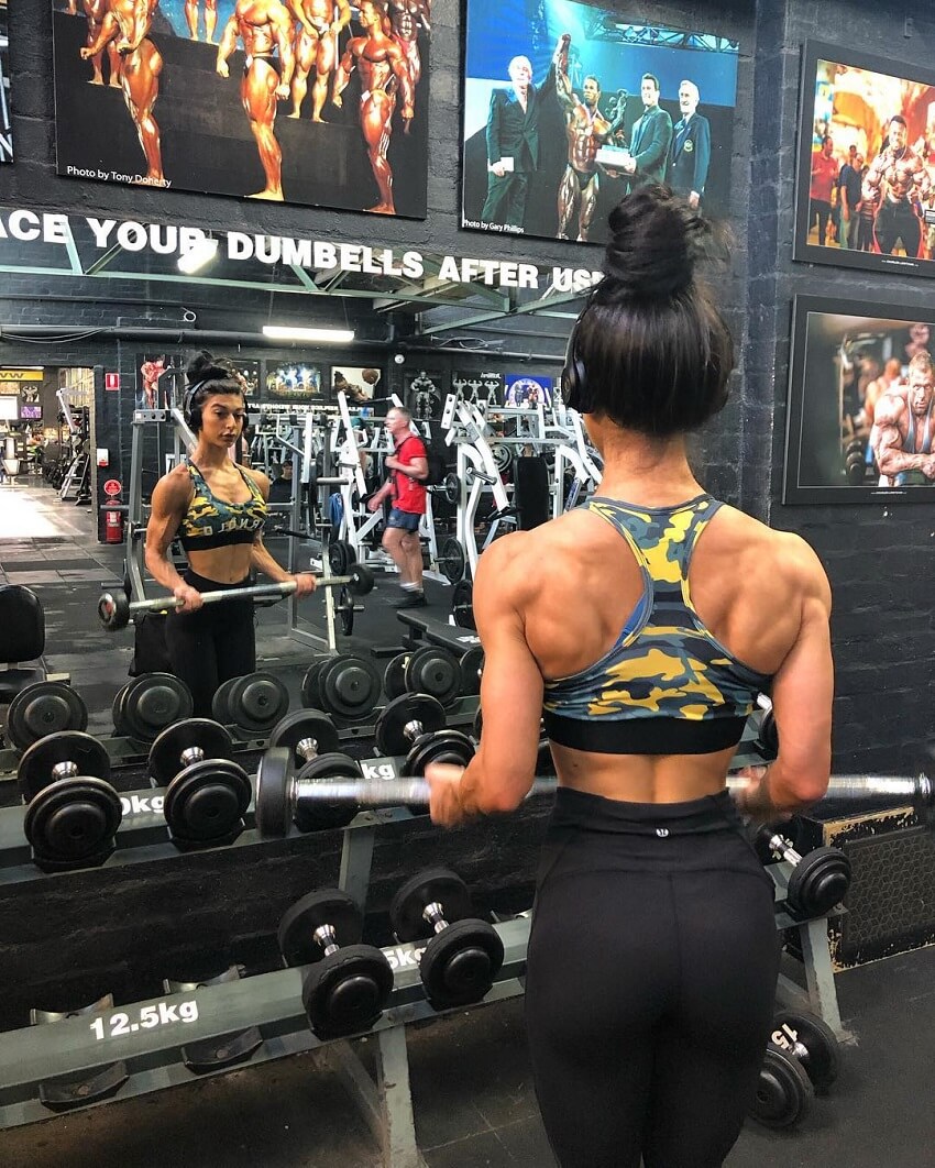 Lauralie Chapados doing barbell bicep curls in front of a dumbbell rack in a gym