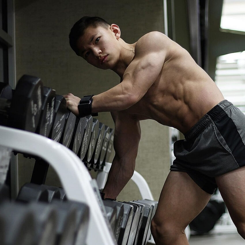 Tristyn Lee posing shirtless next to a dumbbell rack in a gym