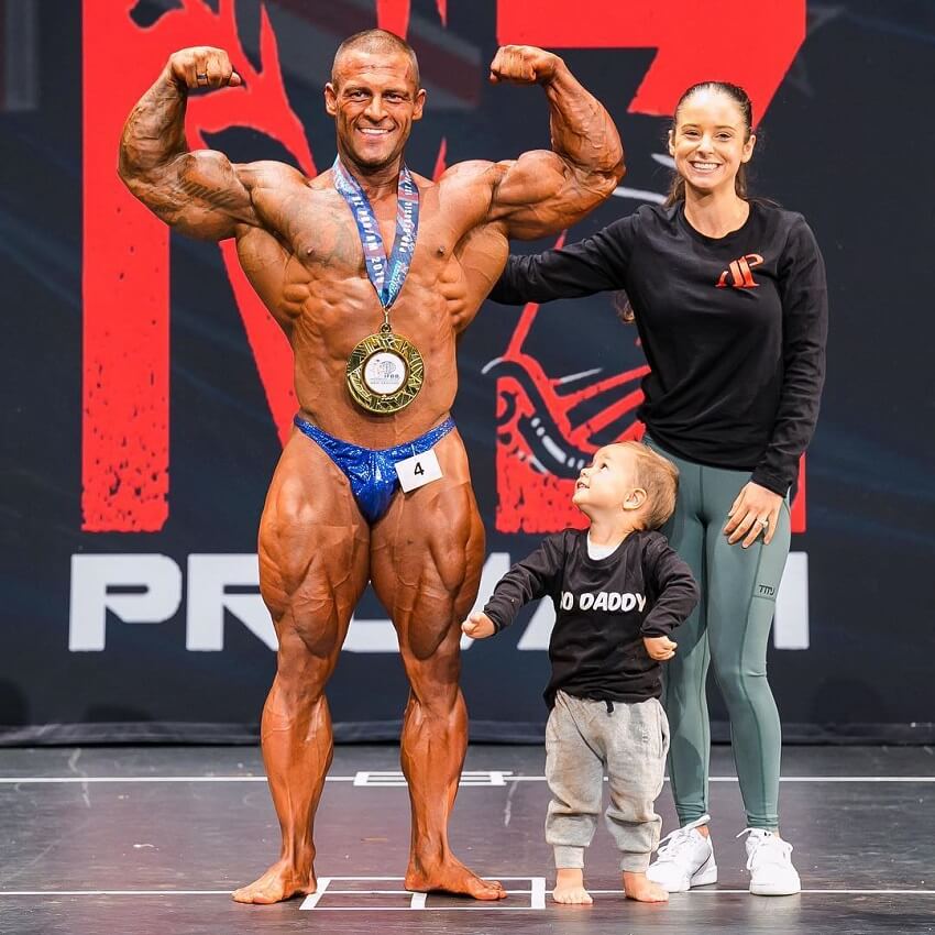 Aaron Polites posing victorious on a bodybuilding stage with his wife and child