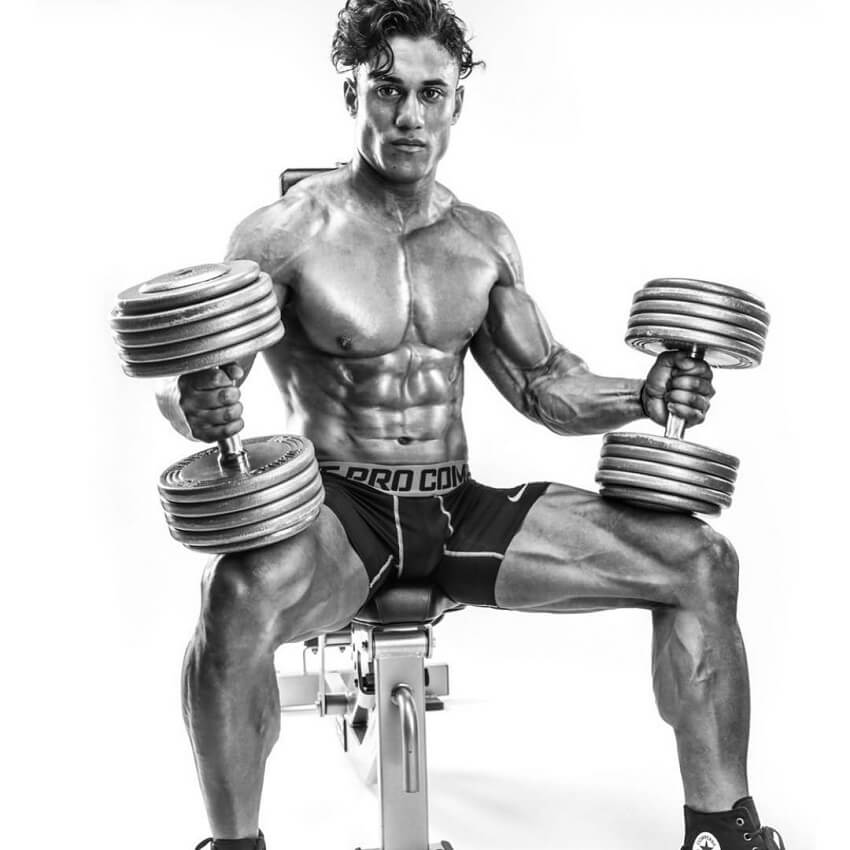 Stephen Pinto sitting shirtless on a bench with dumbbells in his hands