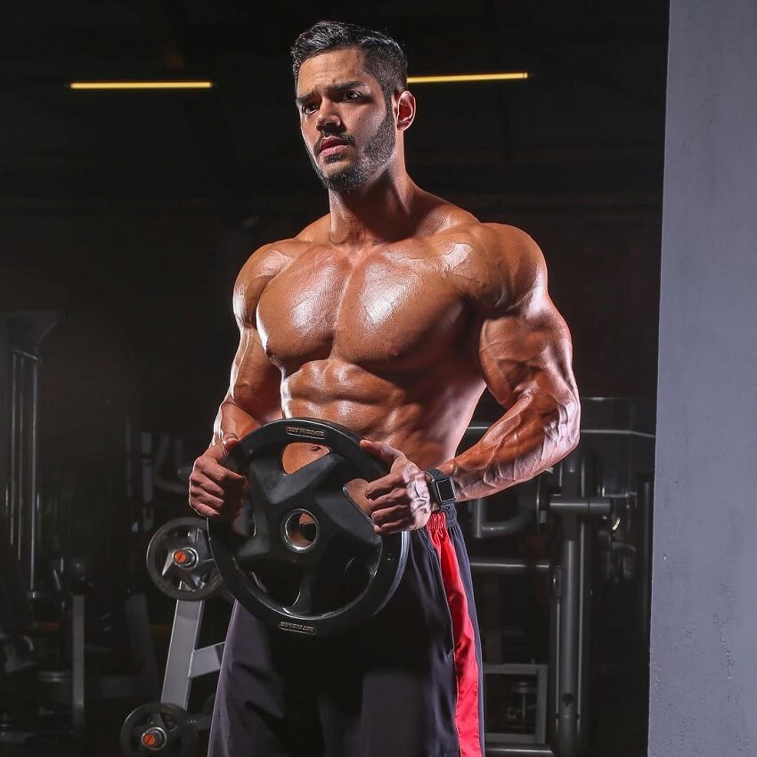 Geder Rocha standing shirtless in the gym, holding a weight plate