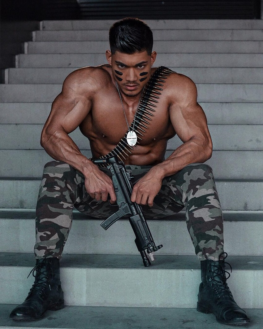 Nicolas Iong sitting shirtless wearing military pants and holding a gun