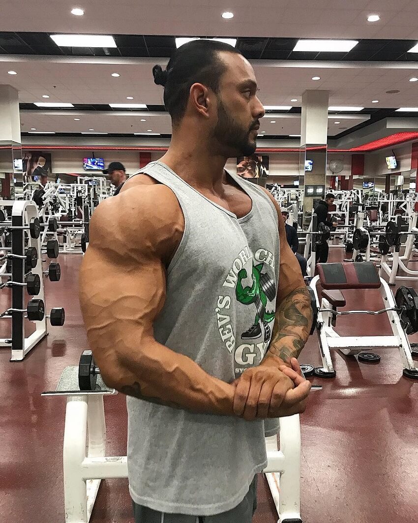 Mark Anthony flexing his ripped and vascular arms in a grey tank top