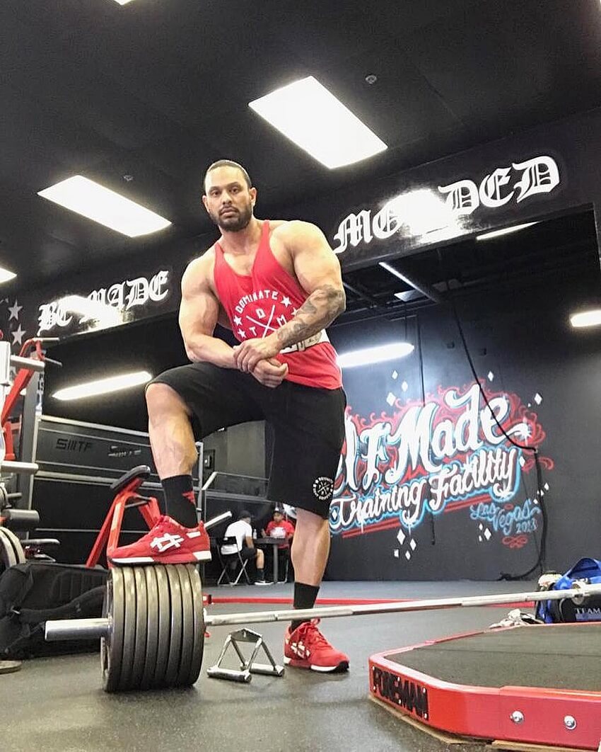 Mark Anthony posing in the gym next to a loaded barbell