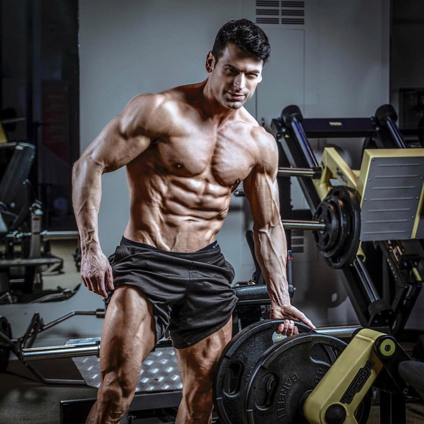 TJ Hoban posing shirtless next to a leg press machine in the gym