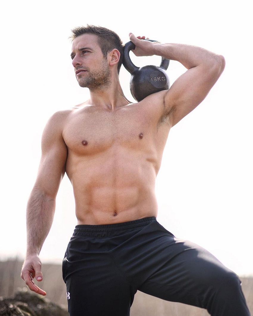 Alex Crockford holding a kettlebell while shirtless outdoors in the sun, looking fit and lean