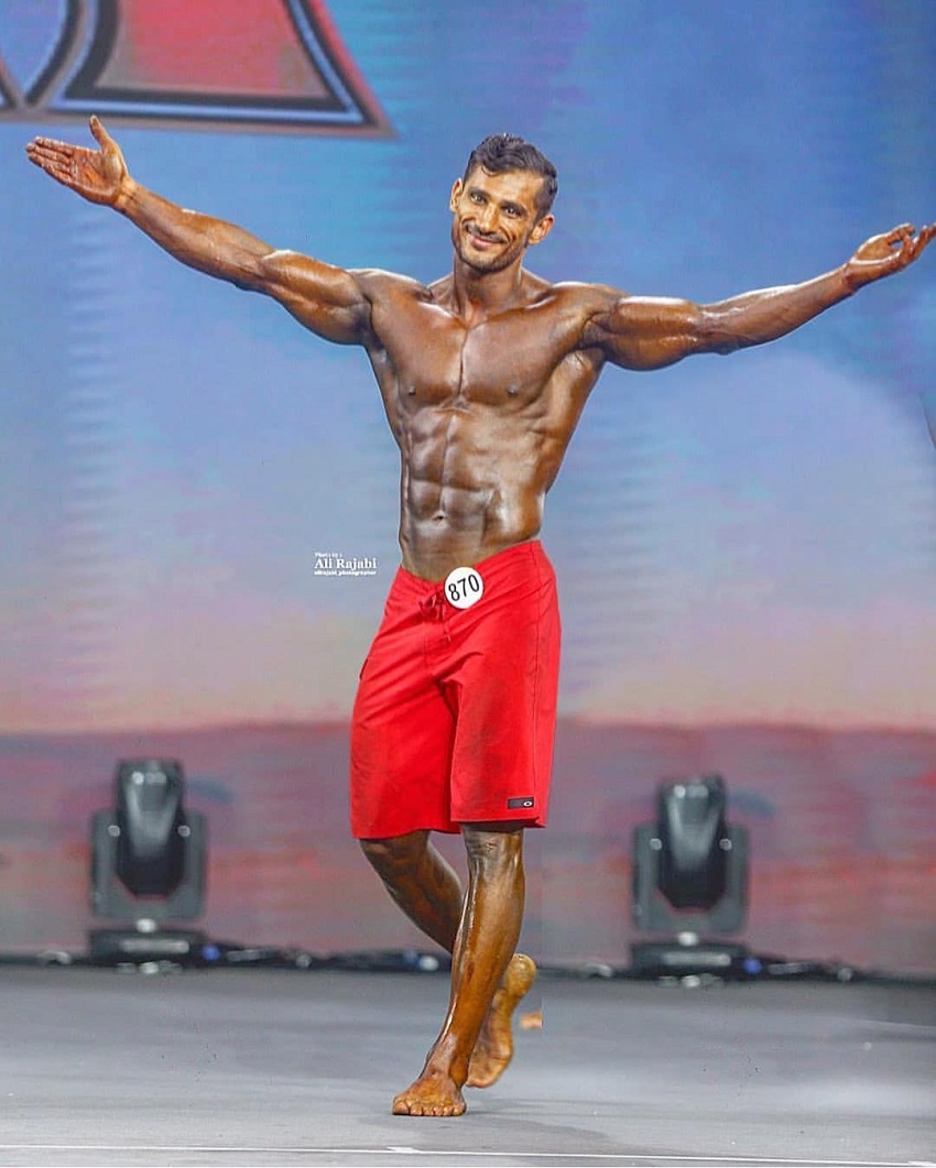 Akbar Sarbaz standing on a bodybuilding stage with his arms spread wide, looking happy with his achievement